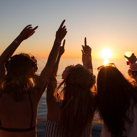 Three women facing a sunset on a boat with their arms in the air