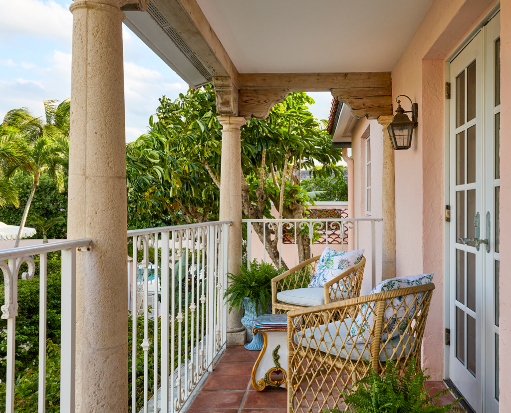 Balcony with two wicker chairs, French doors that lead inside, iron railing around balcony and greenery below on the ground level.