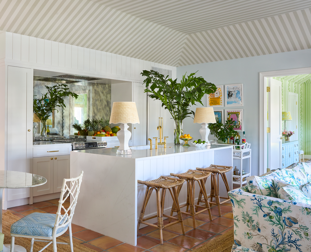 Kitchen with white countertop, blue floral sofa, small bistro table with a chair, palm fronds in vases on the counter, two lamps on the counter and 3 barstools at the counter.