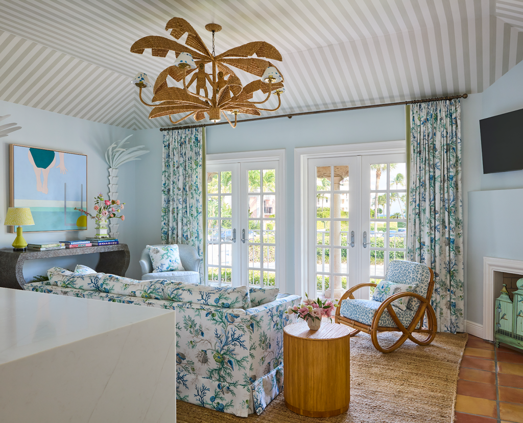 Villa living room, rattan chandelier, round side table,french doors leading to balcony, dark wood console table, floral sofa, wall mounted tv, and fireplace.