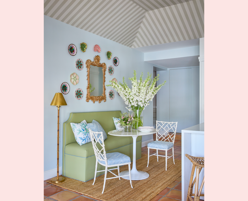 Small bistro table with a green banquette and two white chairs, wall art featuring decorative dishes and a mirror, floral arrangement on table and a tall floor lamp.