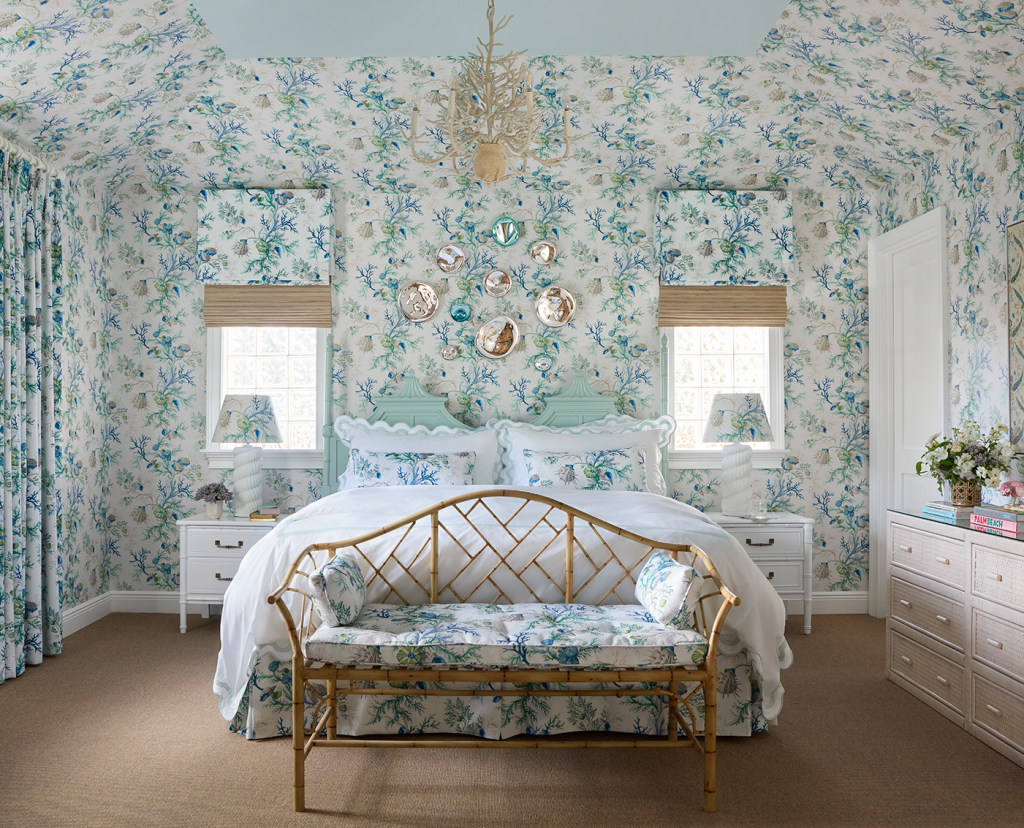 Primary bedroom, blue floral wallpaper on all walls, king bed with matching floral bedding, two windows with brown rattan window treatments, white nightstands on each side of the bed, large white dresser, chandelier, lamps on each nightstand and a rattan bench at the end of the bed.