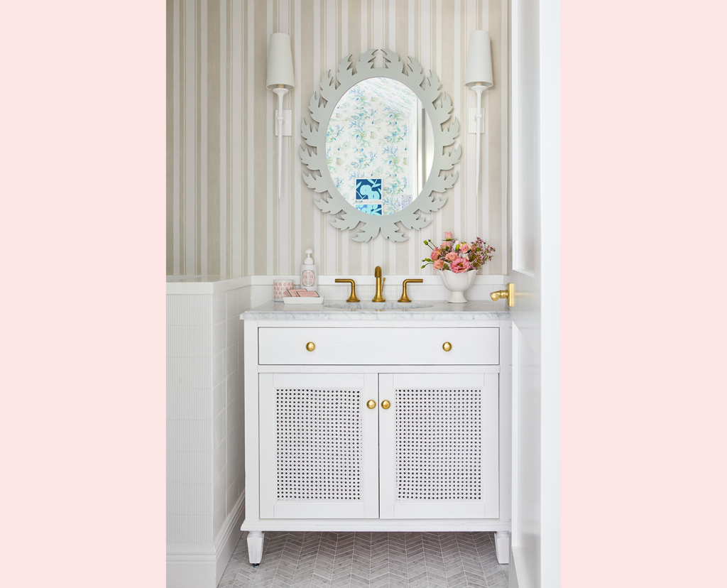 Single vanity bathroom with off-white striped wallpaper, a round white mirror, gold faucet and small wall mounted lamps on each side of the mirror.