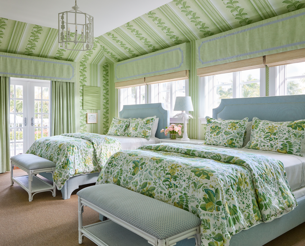 Bedroom with two queen beds, green floral bedding, benches with blue cushions at the end of each bed, Green floral wallpaper, blue headboards and a nightstand in between beds with small white table lamp.