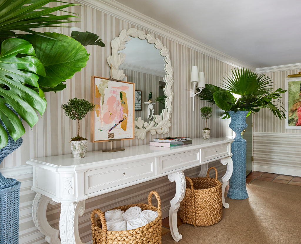 Foyer hallway with white console table, two baskets underneath, flanked by two blue rattan planters with palm fronds in them. White mirror and small artwork on table along with books. Striped off white wallpaper.